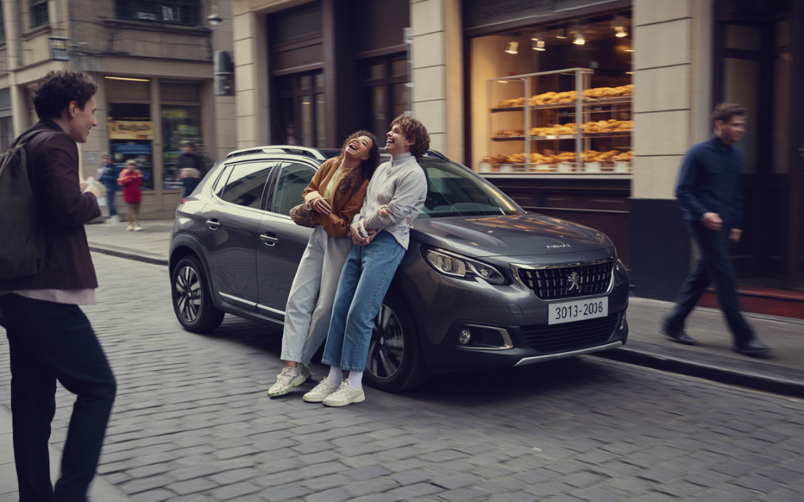 Deux jeunes femmes rient ensemble près d'une voiture grise Peugeot, tandis qu'un homme passe en arrière-plan dans une rue animée.