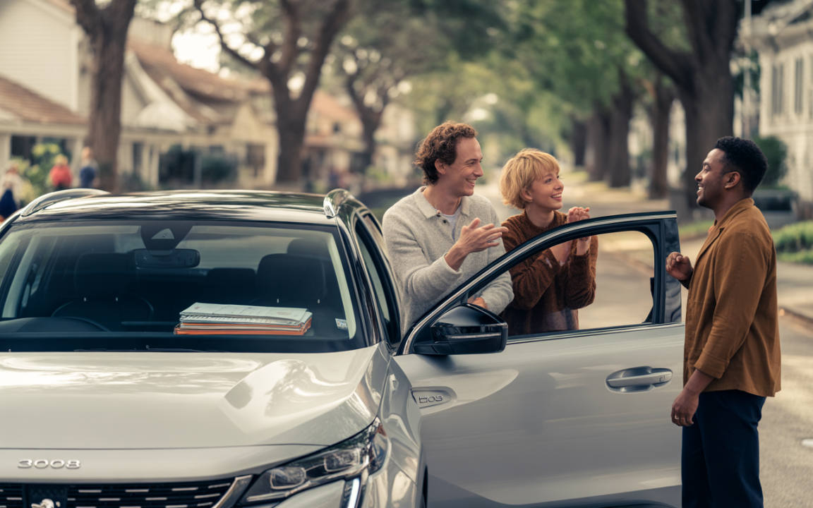 Un couple souriant discute avec un ami près d'une voiture blanche sur une rue bordée d'arbres. Atmosphère amicale et décontractée.