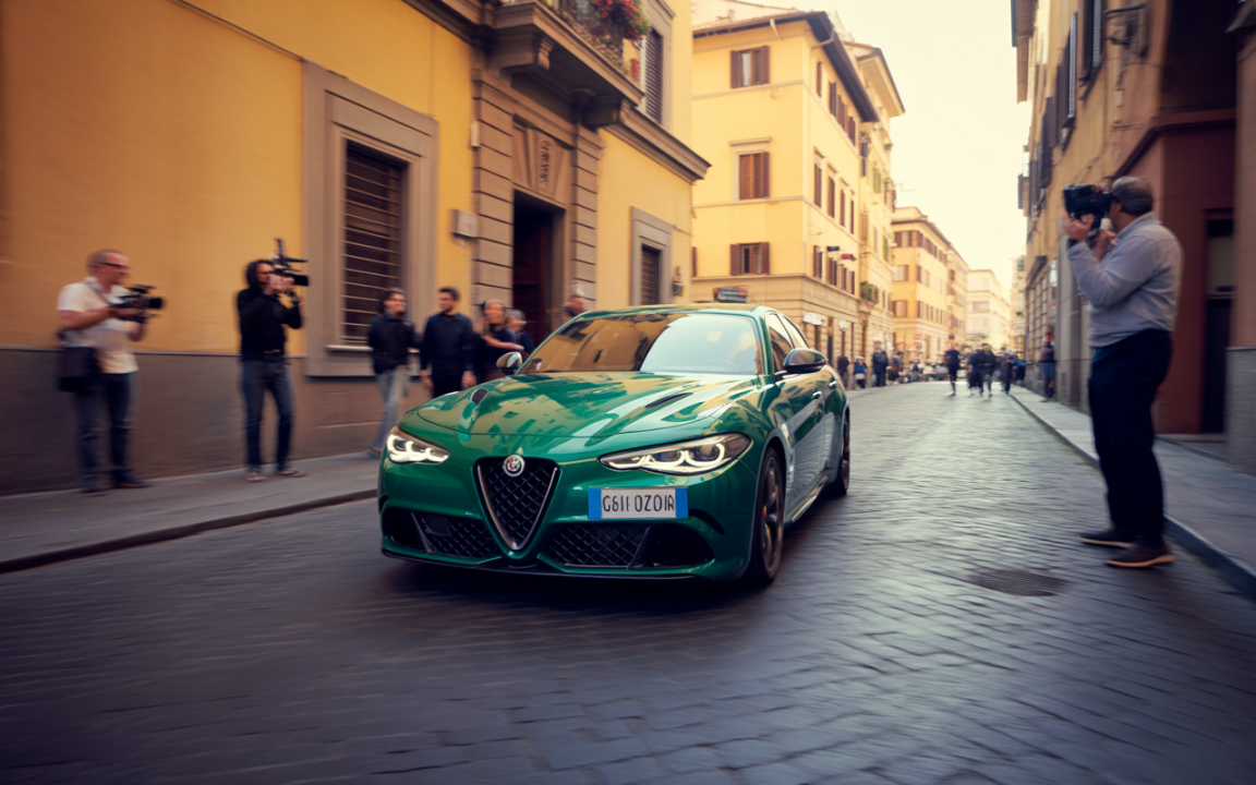 Une voiture verte Alfa Romeo circule dans une rue animée, entourée de photographes et de passants.