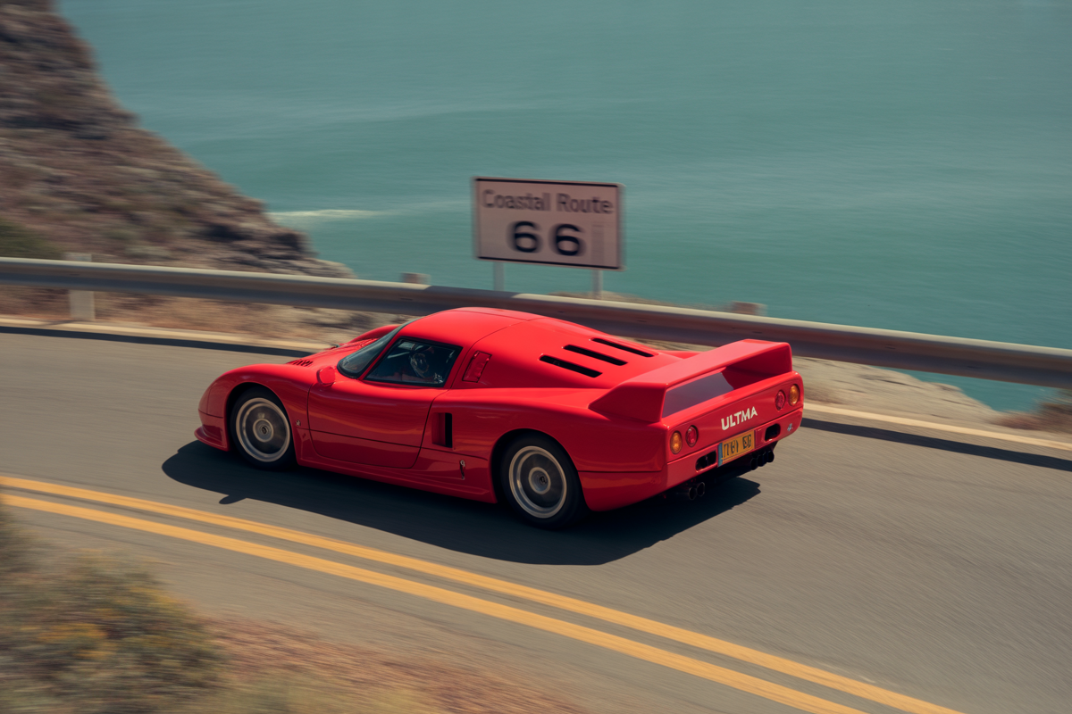 Voiture de sport rouge roulant sur une route côtière, avec un panneau indiquant la Coastal Route 66 en arrière-plan.