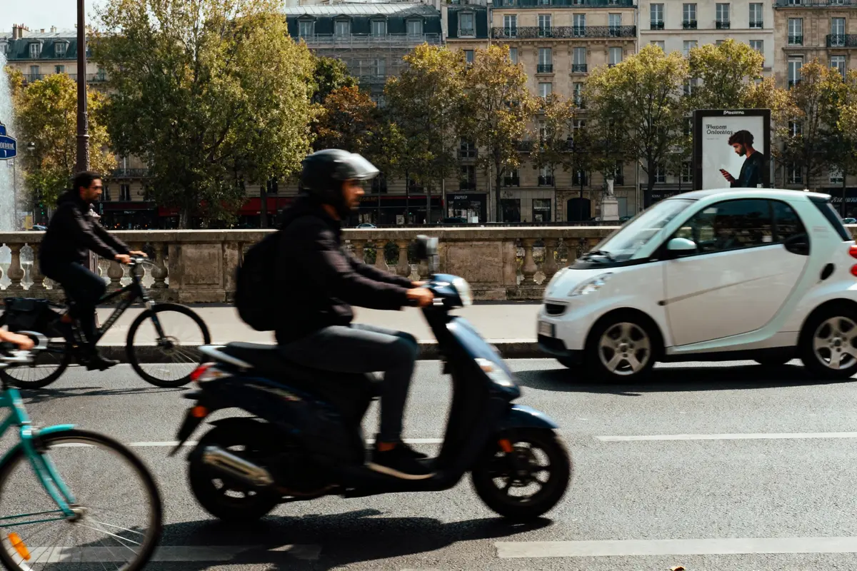 Deux cyclistes, un scooter et une voiture traversant une rue urbaine bordée d'arbres et d'immeubles.