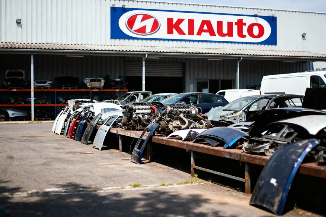 Pièces automobiles et moteurs exposés devant un bâtiment industriel KIAuto, avec voitures à l'arrière-plan.
