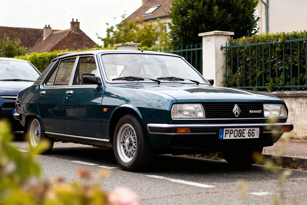 Voiture classique Renault sur route résidentielle, près de maisons et végétation, vue trois-quarts avant.