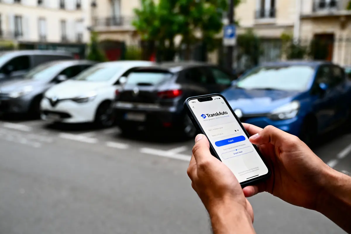 Smartphone utilisant l'application TranskAuto devant des voitures garées dans la rue, flou d'arrière-plan.