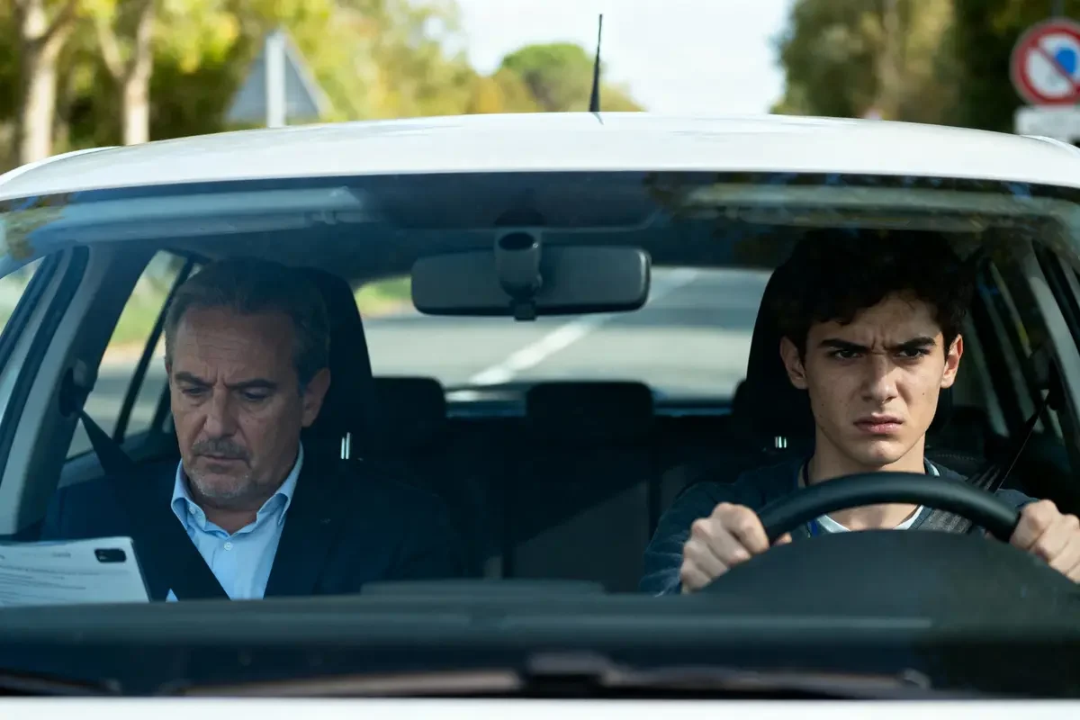 Deux hommes dans une voiture, conducteur concentré, passager lit un document. Route en arrière-plan.