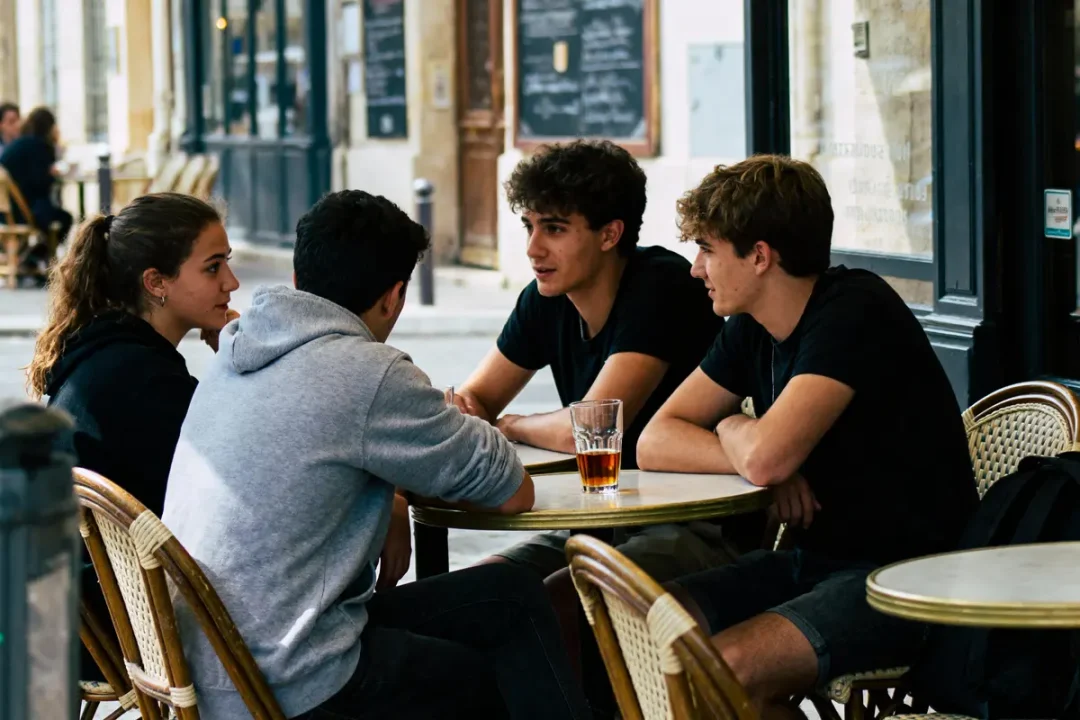 Quatre personnes discutant autour d'une table en terrasse, un verre au centre. Ambiance décontractée de café.