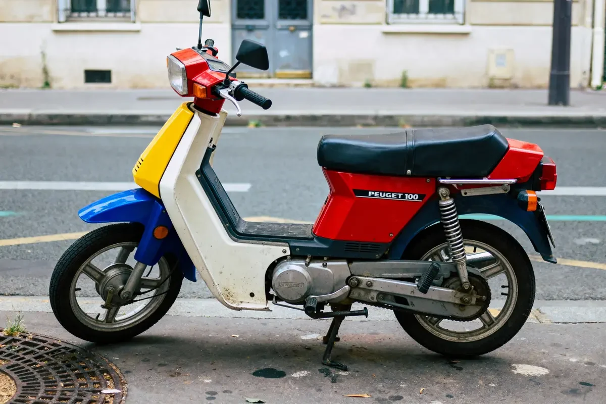 Scooter Peugeot 103 coloré, stationné sur un trottoir, avec carénages rouges, jaunes et bleus, vues urbaines en arrière-plan.
