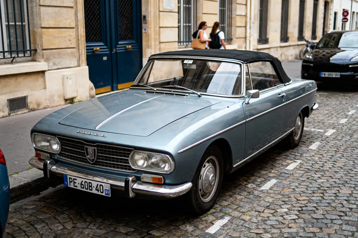 Voiture ancienne bleue cabriolet stationnée sur une rue pavée, avec bâtiments en arrière-plan.