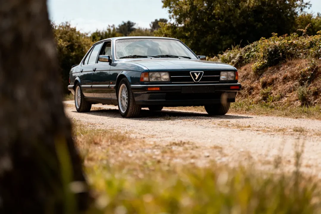 Berline ancienne noire sur chemin de gravier, entourée de végétation dense et ciel dégagé.