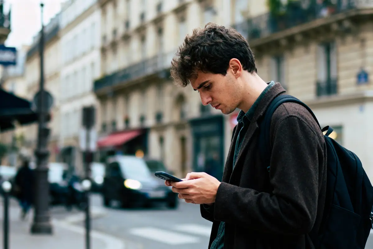 Jeune homme avec un sac à dos regardant son téléphone dans une rue urbaine animée.