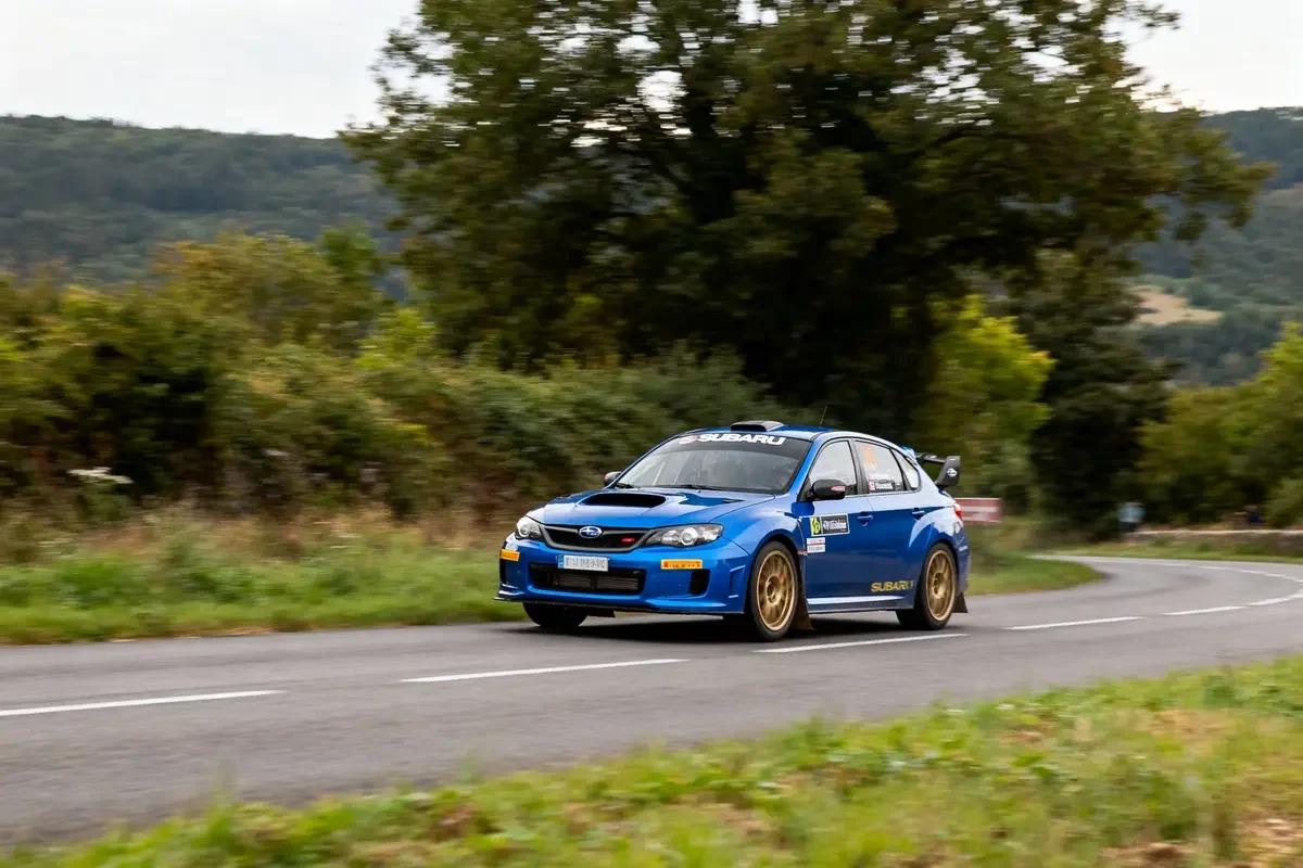 Voiture de rallye bleue Subaru roulant sur route sinueuse entourée de végétation et collines.