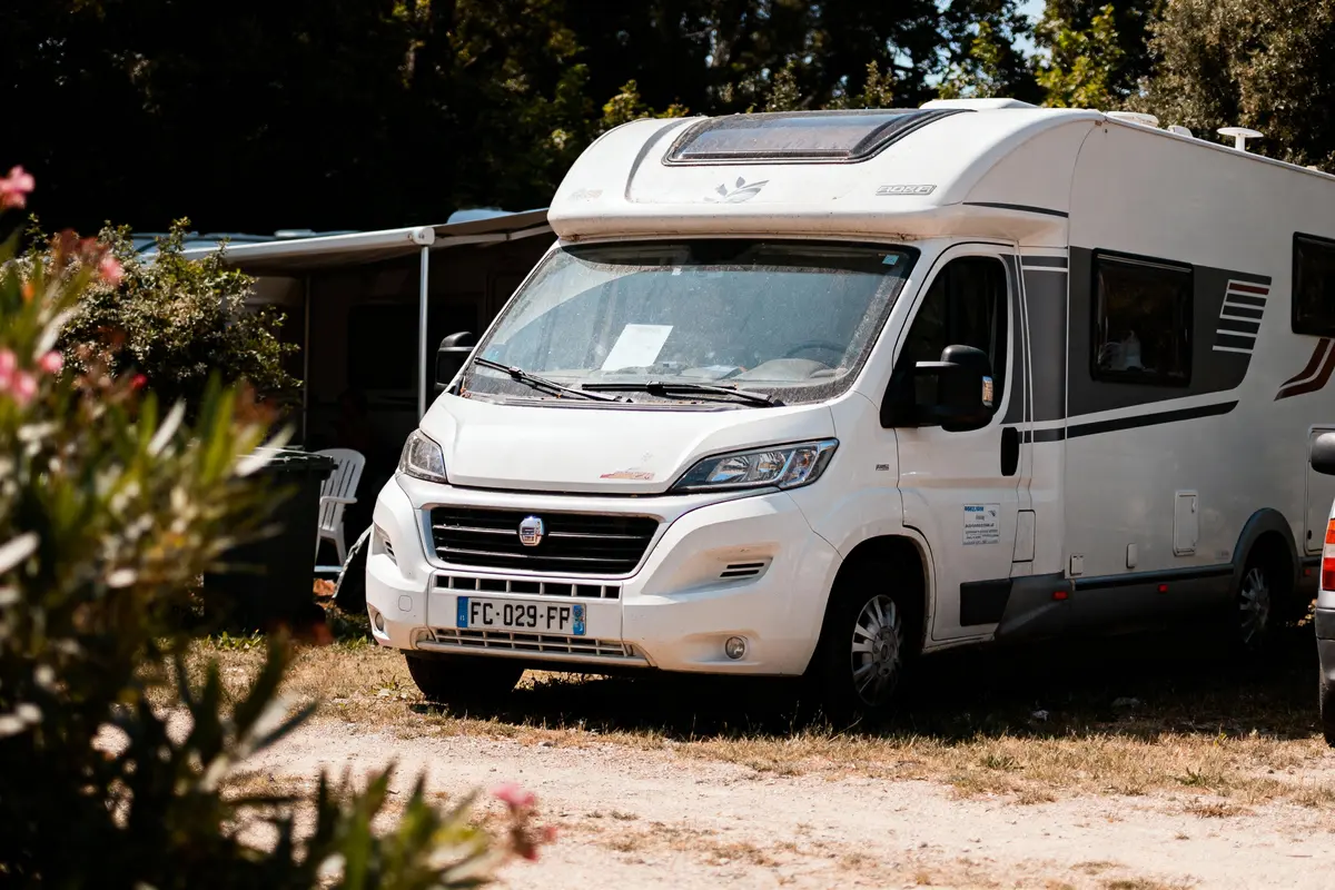 Camping-car blanc garé sur herbe près d'arbustes, arbres en arrière-plan, journée ensoleillée.