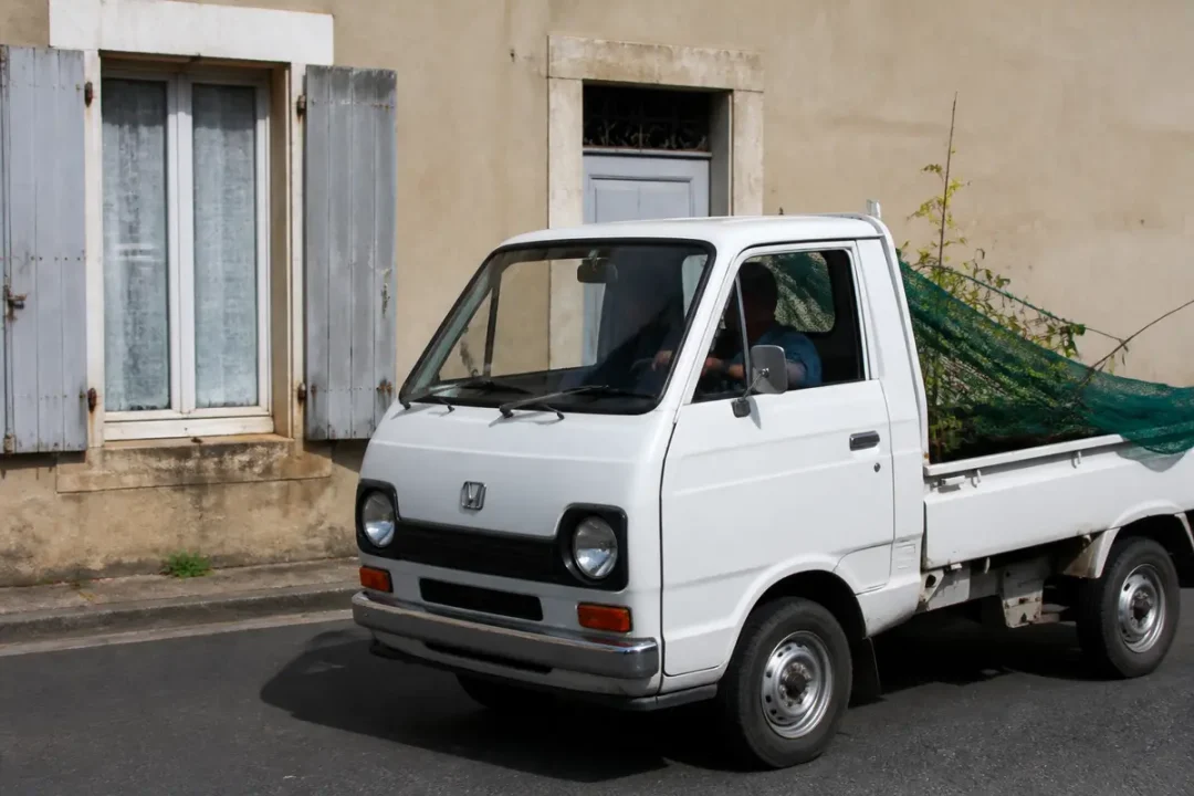 Camionnette blanche Honda Acty transportant des branches devant une maison avec volets gris