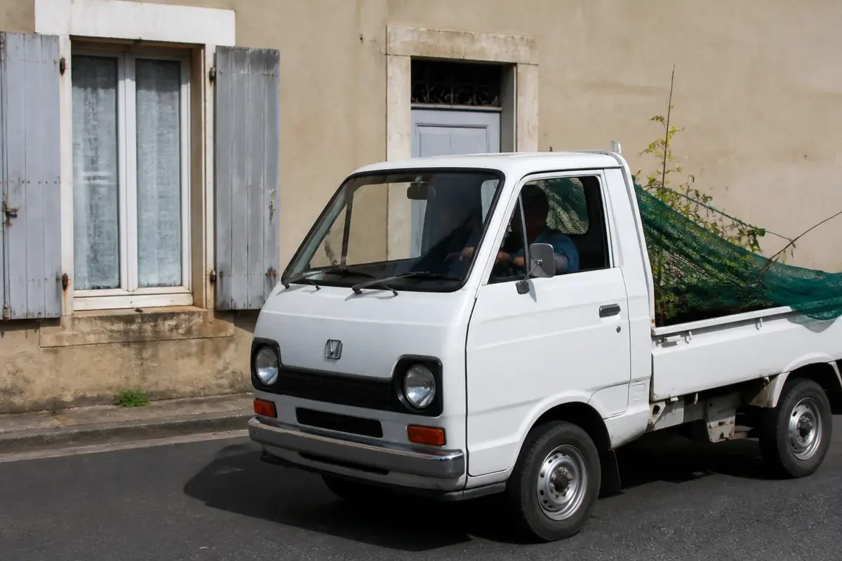 Camionnette blanche Honda Acty transportant des branches devant une maison avec volets gris
