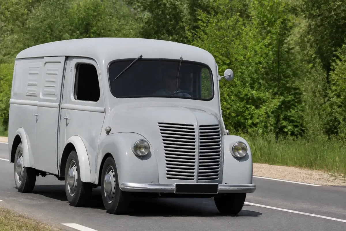 Fourgon ancien Renault Goélette roulant sur route arborée avec pare-brise divisé